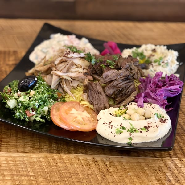 A black plate filled with Middle Eastern food, including shawarma, hummus, tabbouleh, and pickled cabbage.